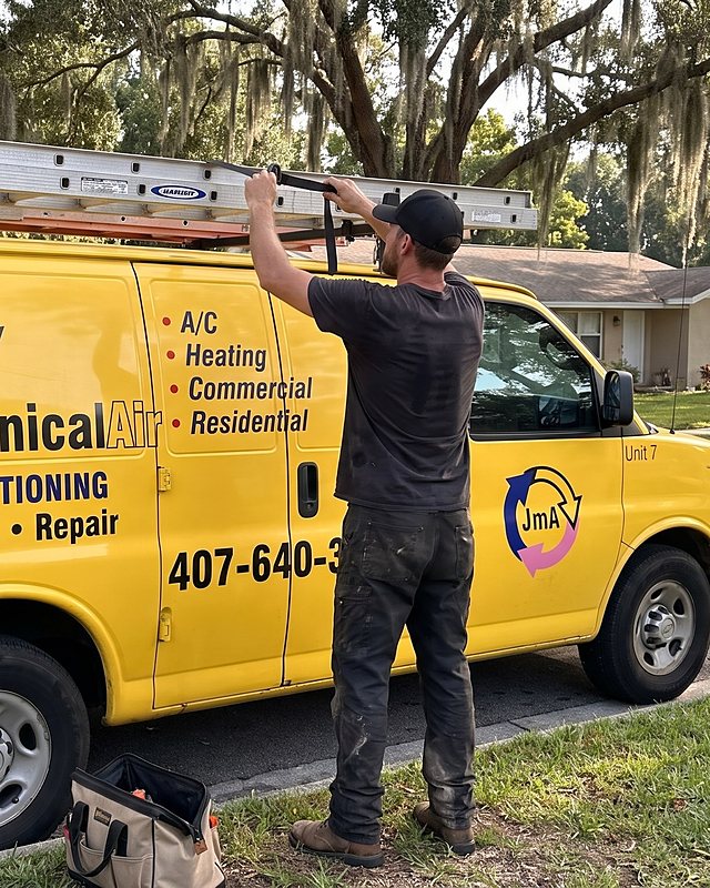 A technician unstraps a ladder from the roof rack of the yellow Jormechanical Air van on an oak-lined street