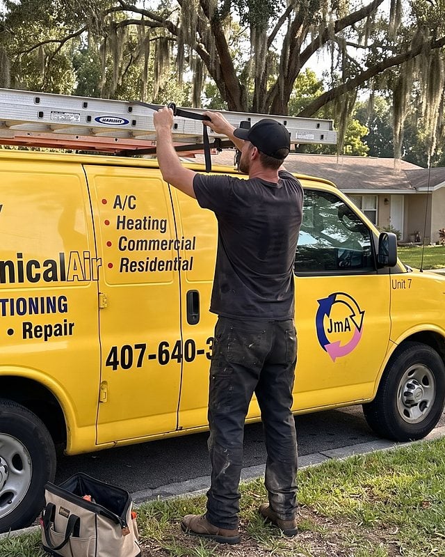 A technician unstraps a ladder from the roof rack of the yellow Jormechanical Air van on an oak-lined street