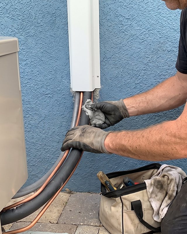 Gloved hands finish the insulation on a refrigerant line set at the wall
