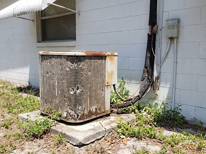 A worn condenser with pitted fins and rotted line set insulation sits on a cracked pad beside a block home