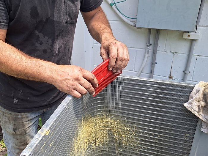 A technician's hands draw a red fin comb along a pollen-dusted condenser coil to straighten bent fins