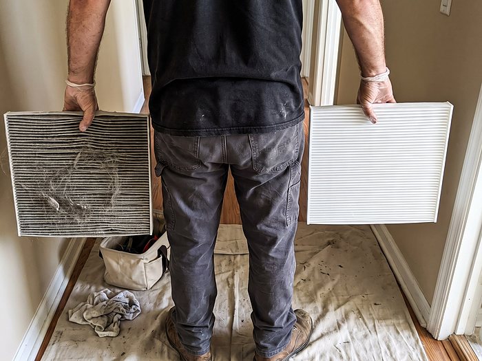 A technician in a hallway holds a clogged, dusty air filter in one hand and a clean white filter in the other