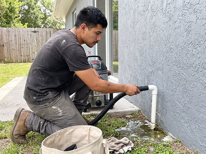 A technician kneels by a stucco wall and vacuums out the white PVC condensate drain line with a wet/dry vac