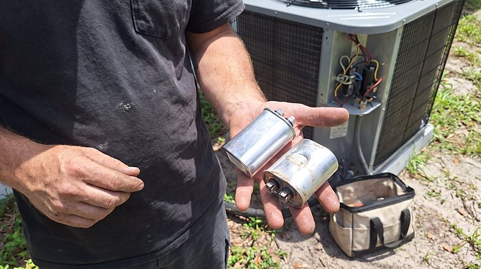 A technician holds a failed capacitor removed from an outdoor condenser