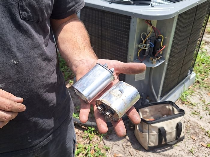 A technician holds a clean capacitor and a rusted, bulging one in his palm beside an open condenser