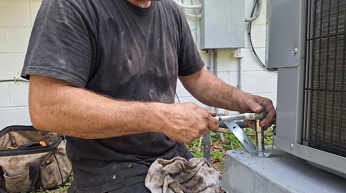 A technician's hands tighten a hurricane tie-down bracket that anchors a condenser to its concrete pad