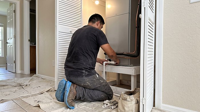 A technician kneels on a drop cloth to work on an air handler in a hallway closet