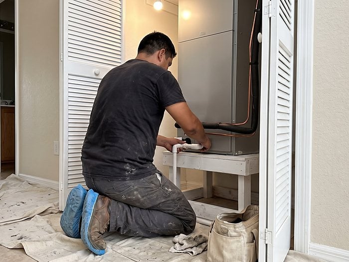 A technician in shoe covers kneels on a drop cloth fitting a PVC drain to an air handler in a hall closet