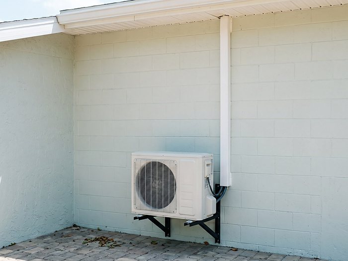 A ductless mini-split outdoor unit on black wall brackets, its white line cover running up a block wall to the eave