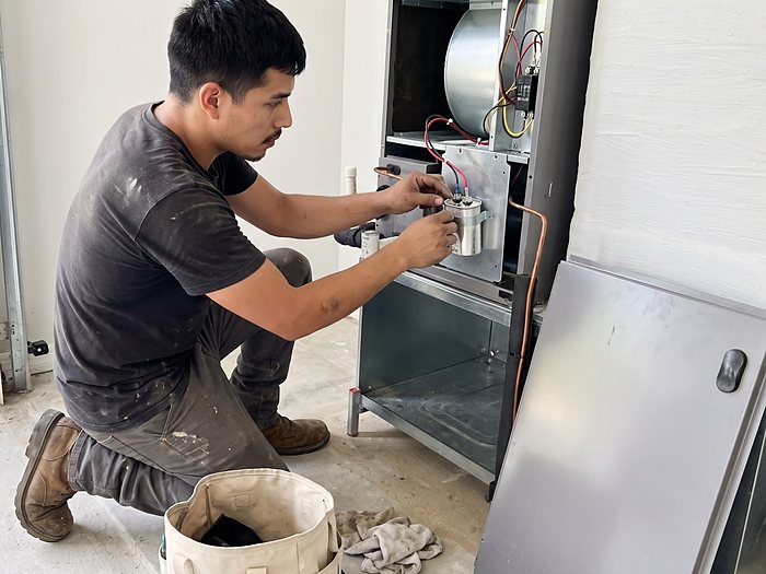 A technician fits a new capacitor into an open air handler in a garage, its front panel leaning nearby