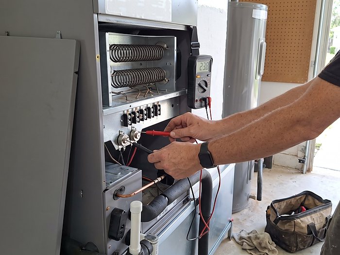 A technician tests the electric heat strip terminals inside an air handler with a multimeter in a garage