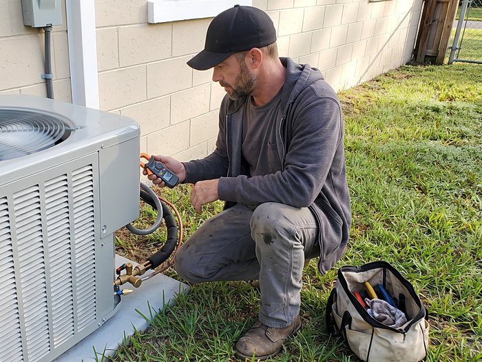 A technician in a hoodie crouches beside a running outdoor unit and reads a clamp meter by the service valves