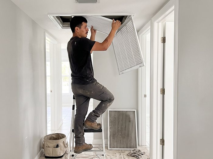 A technician on a step ladder opens a ceiling return air grille in a hallway, a dusty old filter below