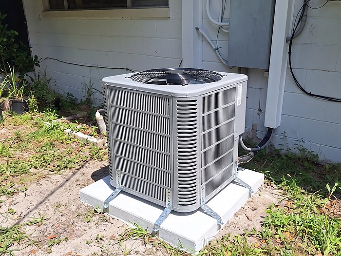 A new outdoor unit on a white pad with metal tie-down straps beside a painted block home and grassy yard