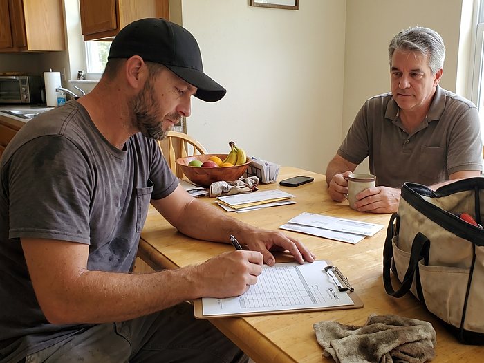 A technician writes up an estimate on a clipboard at a kitchen table while a homeowner holds a coffee mug