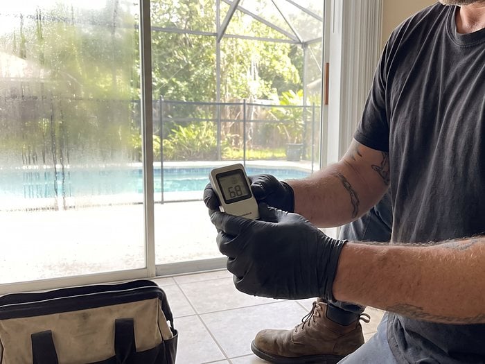 A gloved technician holds a humidity meter reading 68 by a fogged sliding door facing a screened pool