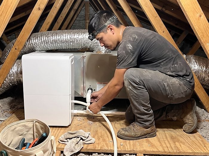 A technician wearing a headlamp connects a drain line to a whole-home dehumidifier beside an attic air handler