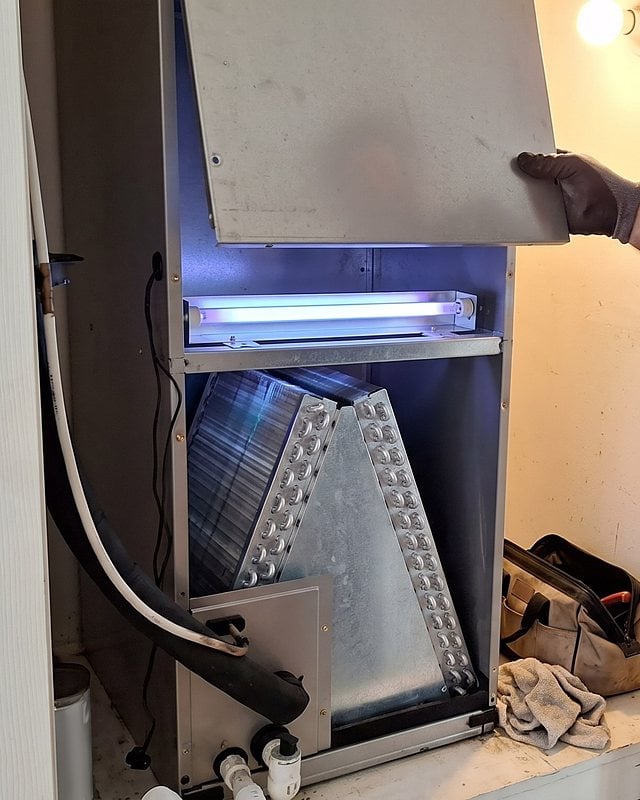 A technician lifts the access panel off an indoor air handler to reveal a glowing UV lamp above the coil