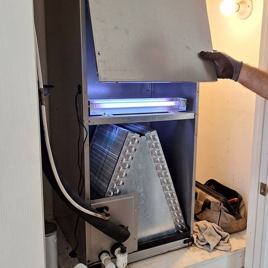 A technician lifts the access panel off an indoor air handler to reveal a glowing UV lamp above the coil