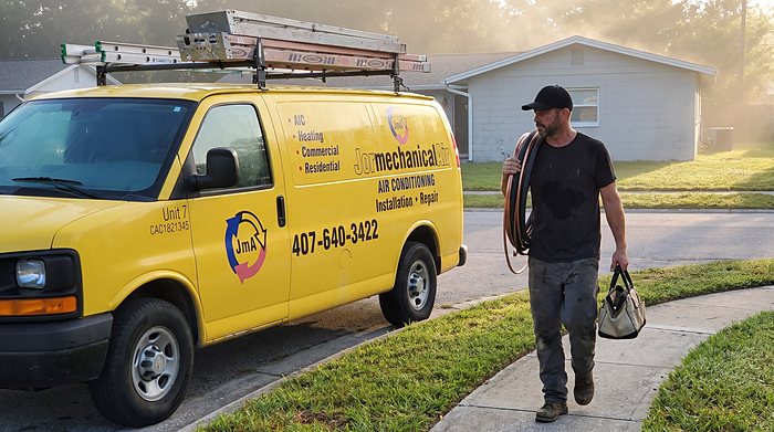 A technician carries a coiled line set and a tool bag from the yellow Jormechanical Air van at sunrise