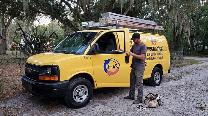 A technician writes on a clipboard beside the yellow Jormechanical Air van under live oaks hung with moss