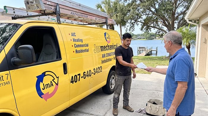 A technician hands paperwork to a homeowner beside the yellow Jormechanical Air van on a lakefront driveway