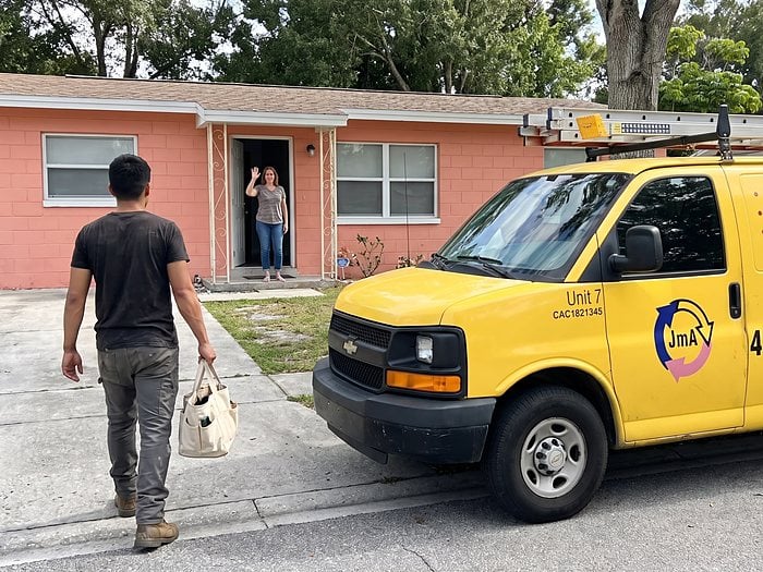 A homeowner waves from her door as a technician walks up from the yellow Jormechanical Air van