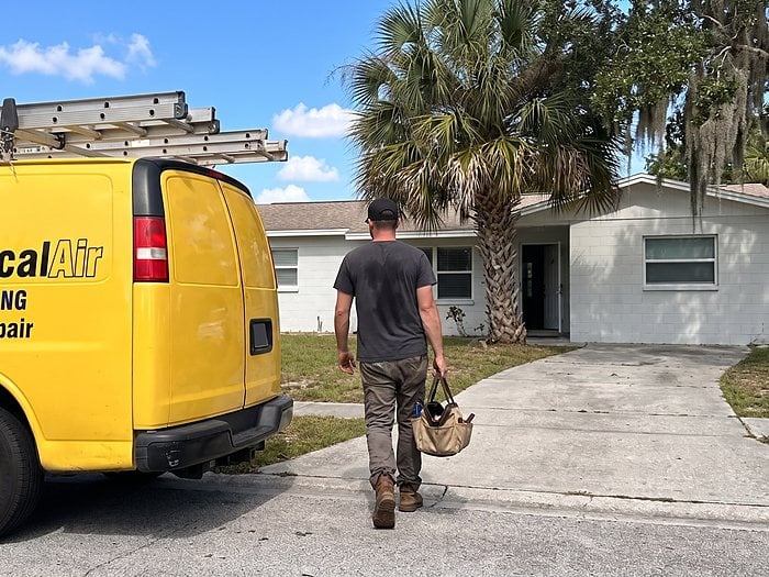 A technician carries a tool bag toward a white block home past the yellow Jormechanical Air van